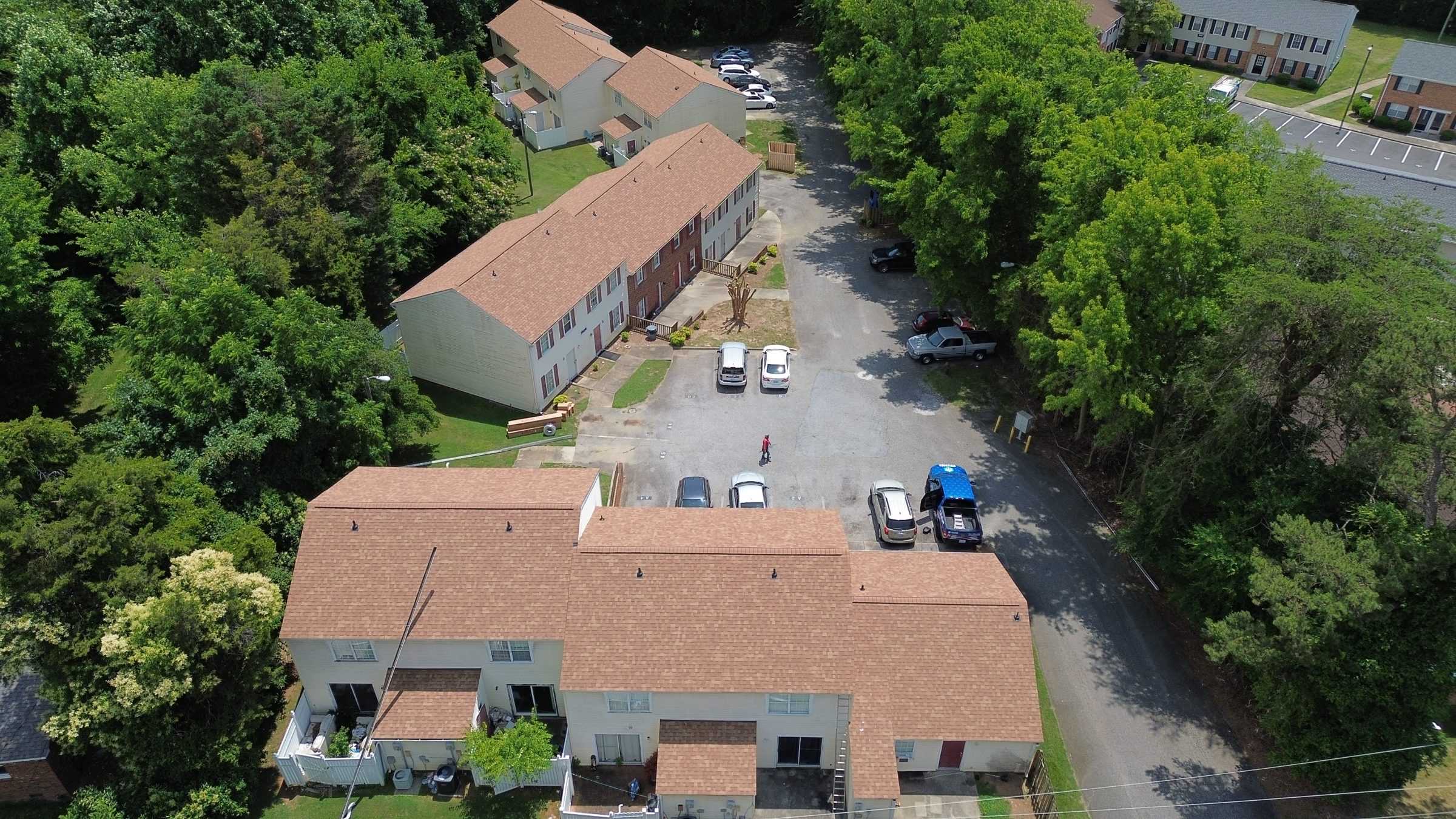 Drone view of two apartment buildings with brown shingle roofs beside a parking lot.