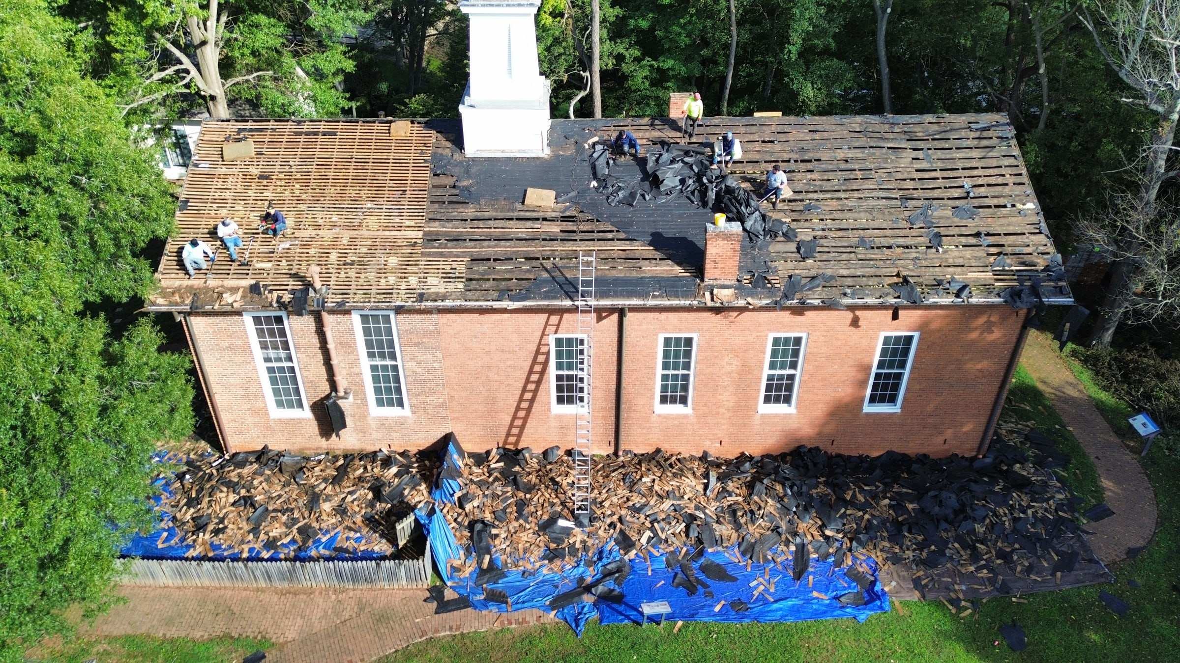 A crew stripping old shingles down to the boards on a brick building, with tarps catching the debris below.